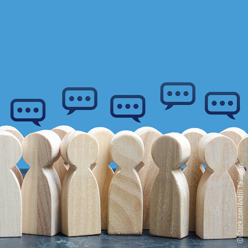 Zahlreiche Holzfiguren stehen vor einem blauen Hintergrund. Über ihren Köpfen schweben Comic-Sprechblasen mit stilisiertem Text.